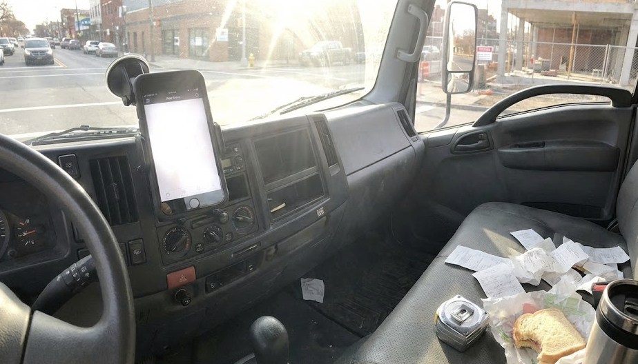 Truck cab interior with phone mounted on dash washed out by sun glare, receipts and food scattered on passenger seat